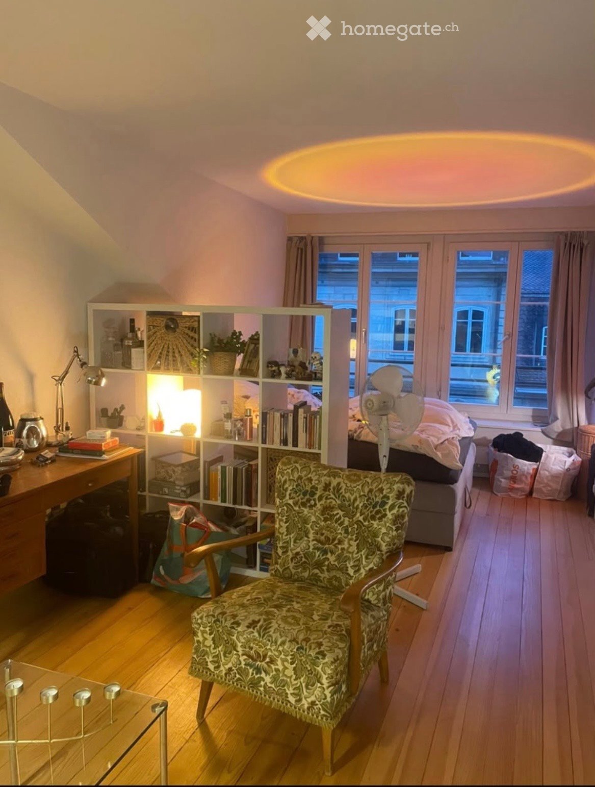 A cozy, well-lit room with wooden flooring, a patterned armchair, a glass coffee table, a bookshelf filled with books and decorative items, a desk with a lamp, and a bed with pillows and a fan nearby.