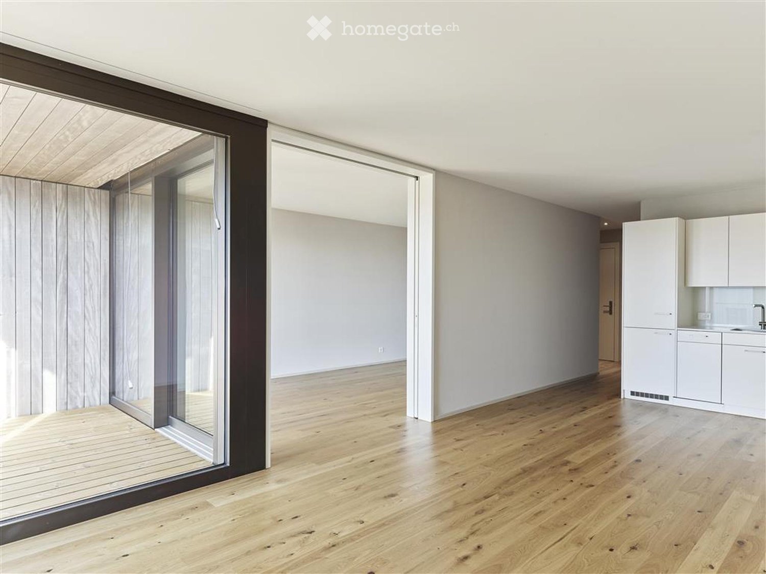Empty room with light wooden flooring, white walls, and a glass door leading to an outdoor area. A small kitchen area is visible in the background with white cabinets and a refrigerator.