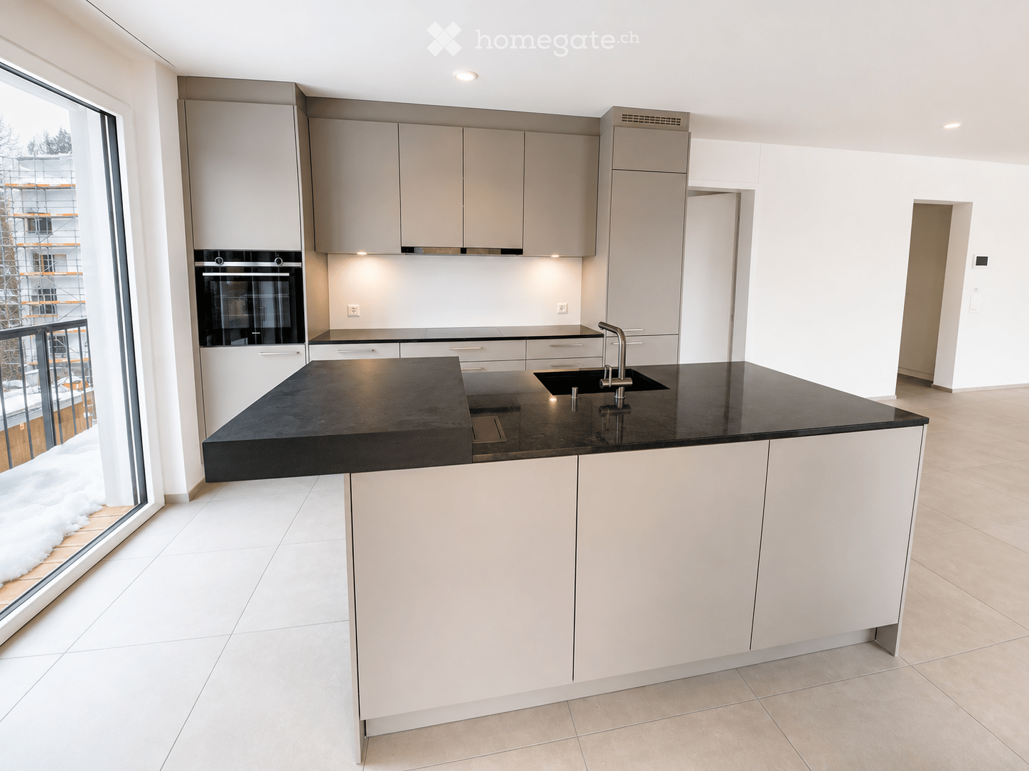 Modern kitchen with white cabinets, black countertop, built-in oven, and a sink with a modern faucet. Large window offers a view of the balcony.