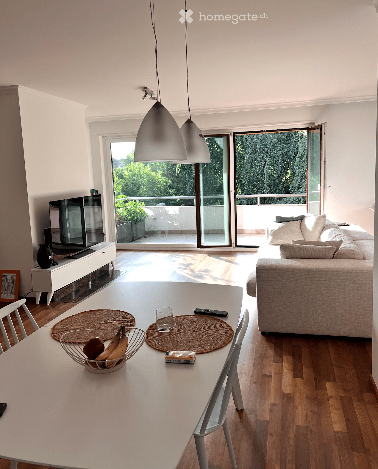 Bright living and dining area with a sofa, dining table, chairs, TV, and pendant lights. Large sliding glass doors open to a balcony with outdoor views.
