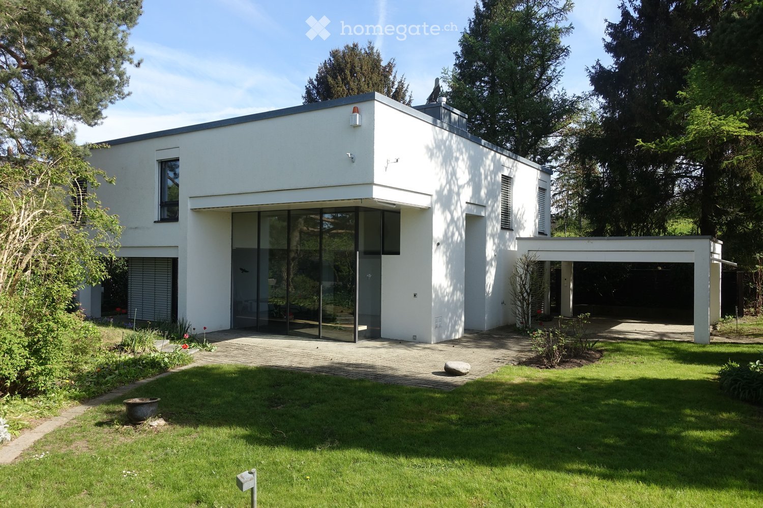Modern two-story house with white exterior, large glass entrance, garage, and well-maintained garden.
