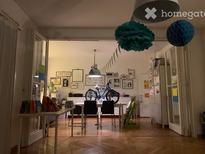 A modern, open-plan living and dining area with wooden flooring, white walls, and large windows. The space features a dining table with chairs, a bicycle, a small cabinet, and several picture frames on the wall. Colorful balloons are hanging from the ceil