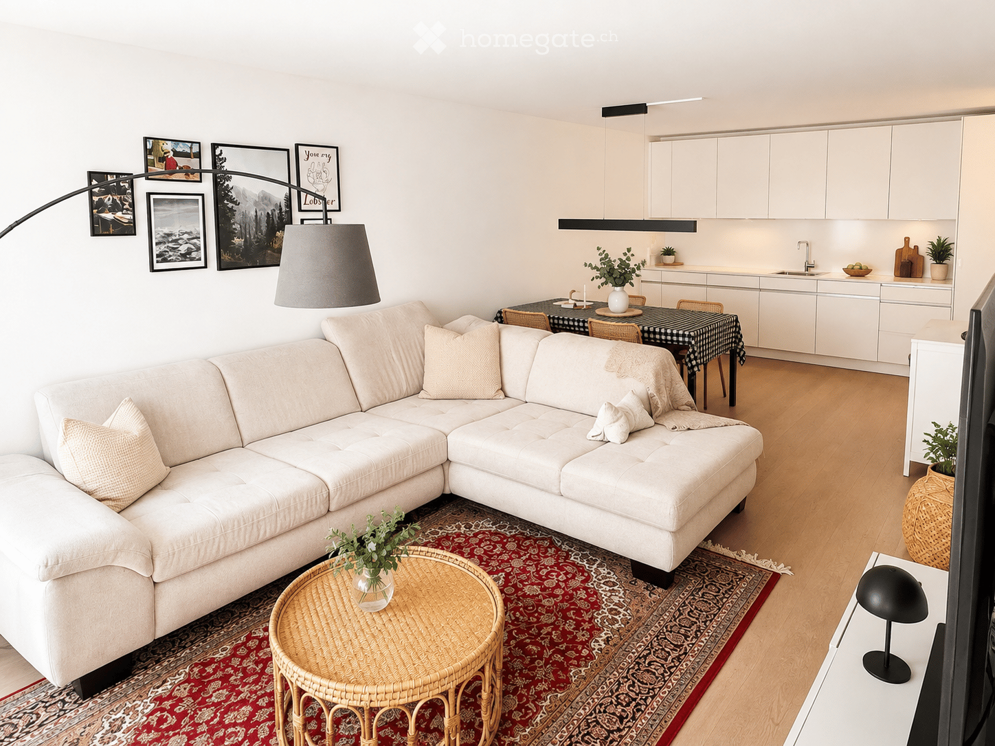 Modern living room with a white sectional sofa, coffee table, dining area with a table and chairs, and a fitted kitchen with white cabinets and a sink.