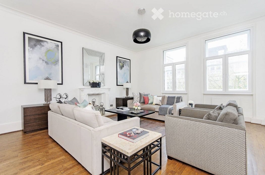 Bright living room with white walls, wooden flooring, and large windows. Features a white sofa, grey armchair, coffee table, side table, fireplace, and decorative items.