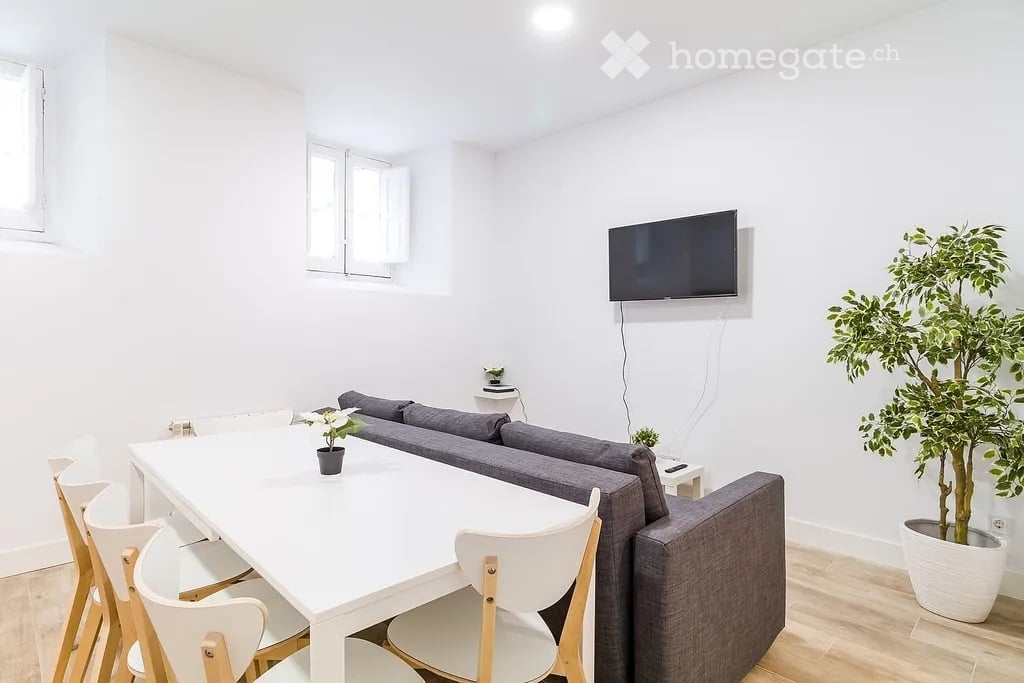 Bright living room with white walls, grey sofa, white dining table with chairs, and a mounted TV. Potted plants are placed on the floor and table.