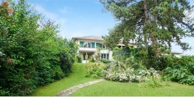 A two-story white house with green shutters, surrounded by lush greenery and a well-maintained garden.