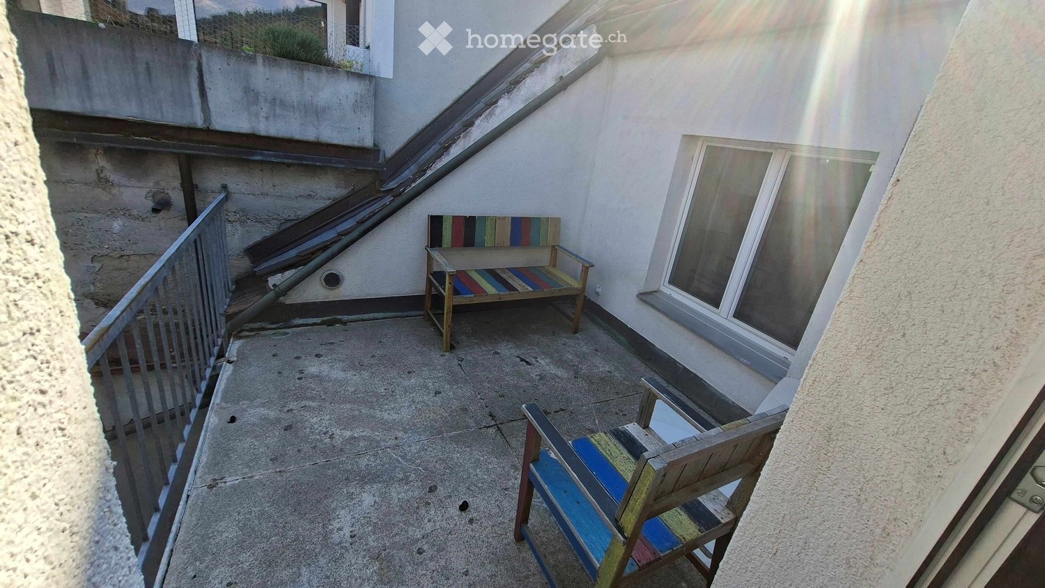 Balcony with a bench and a chair, concrete floor, metal railing, and a window with a view.