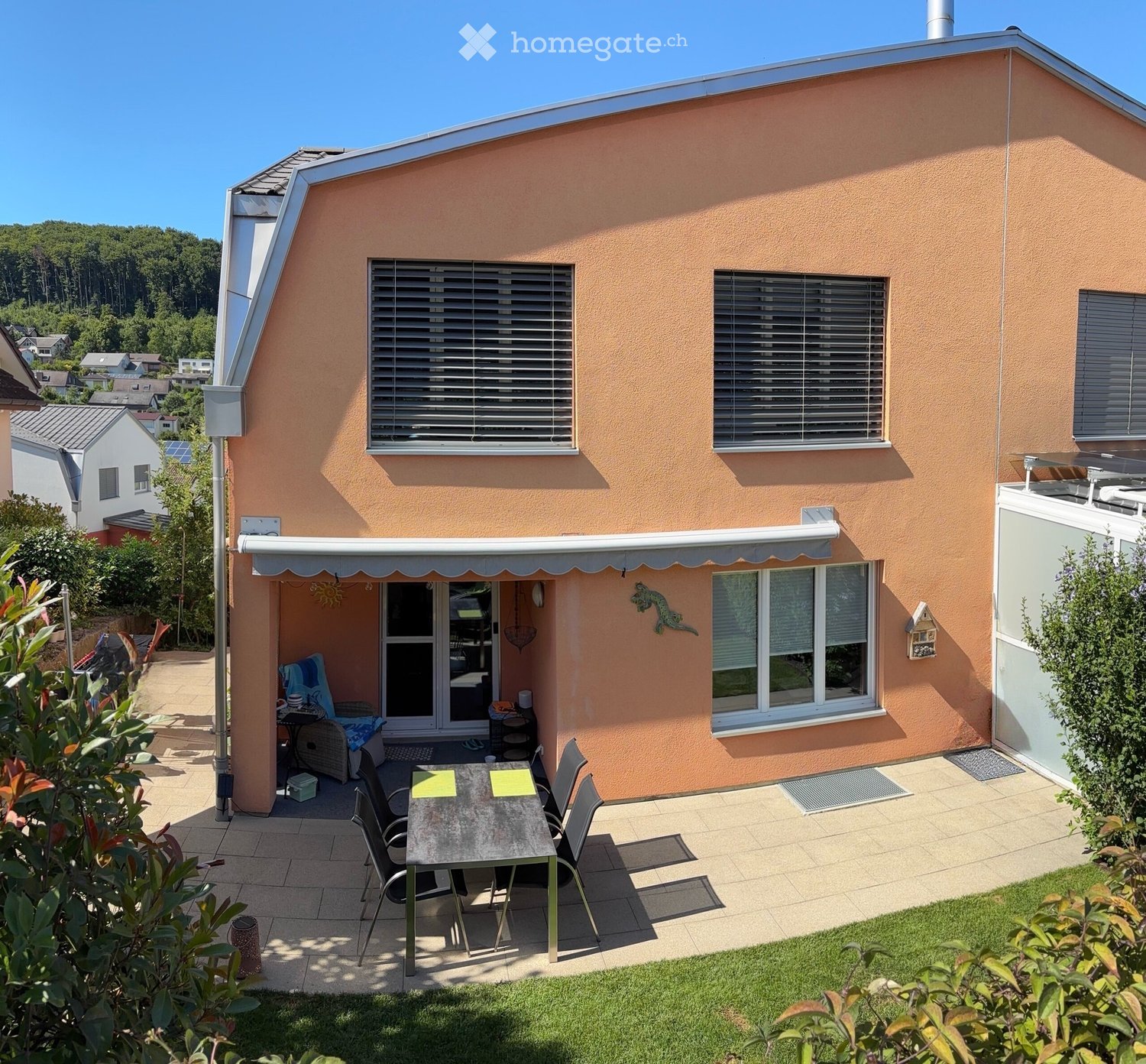 Outdoor patio area with a table, chairs, and an umbrella attached to the house. The house has a peach-colored exterior with multiple windows and a small covered entrance.