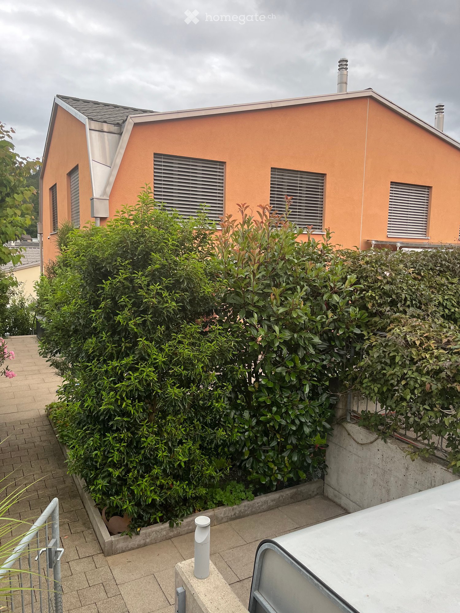 A modern orange house with a flat roof and multiple windows, partially obscured by a dense hedge. The surrounding area includes a paved walkway and a small garden bed.