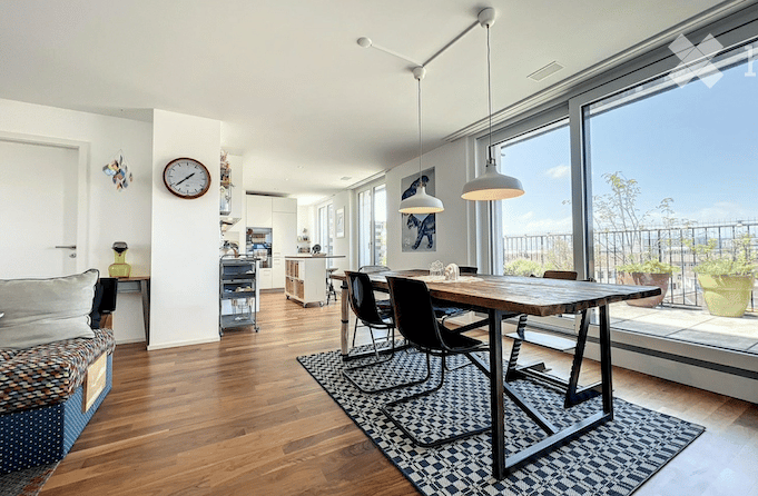 Open-plan living and dining area with hardwood floors, large windows, and a balcony view. Features a sofa, coffee table, dining table with chairs, and a kitchen island.