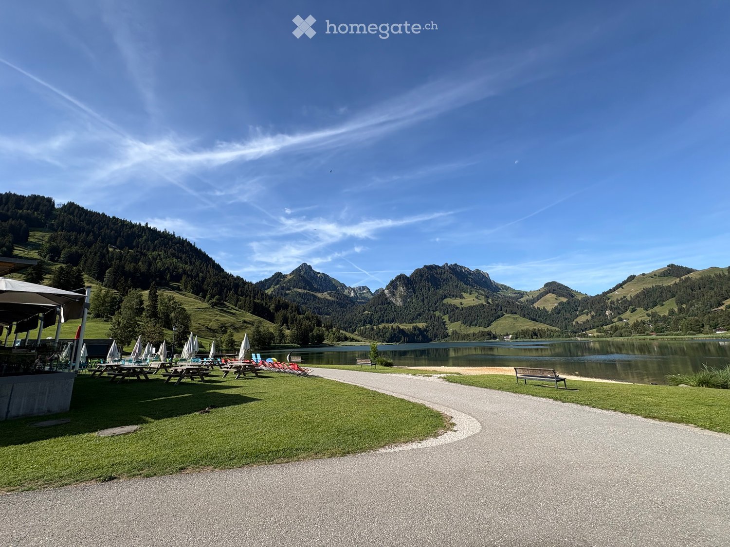 Lakeside park with picnic tables, benches, umbrellas, and lounge chairs, surrounded by green hills and mountains.