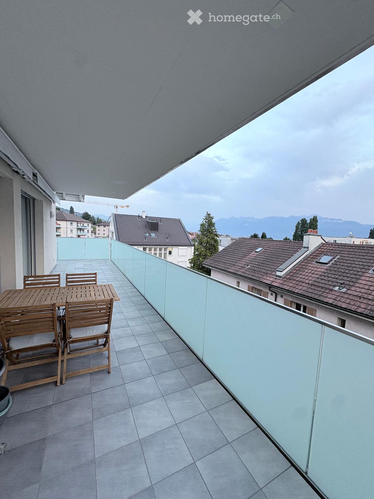 Balcony with tiled floor, glass railing, wooden table and chairs, overlooking cityscape and mountains.