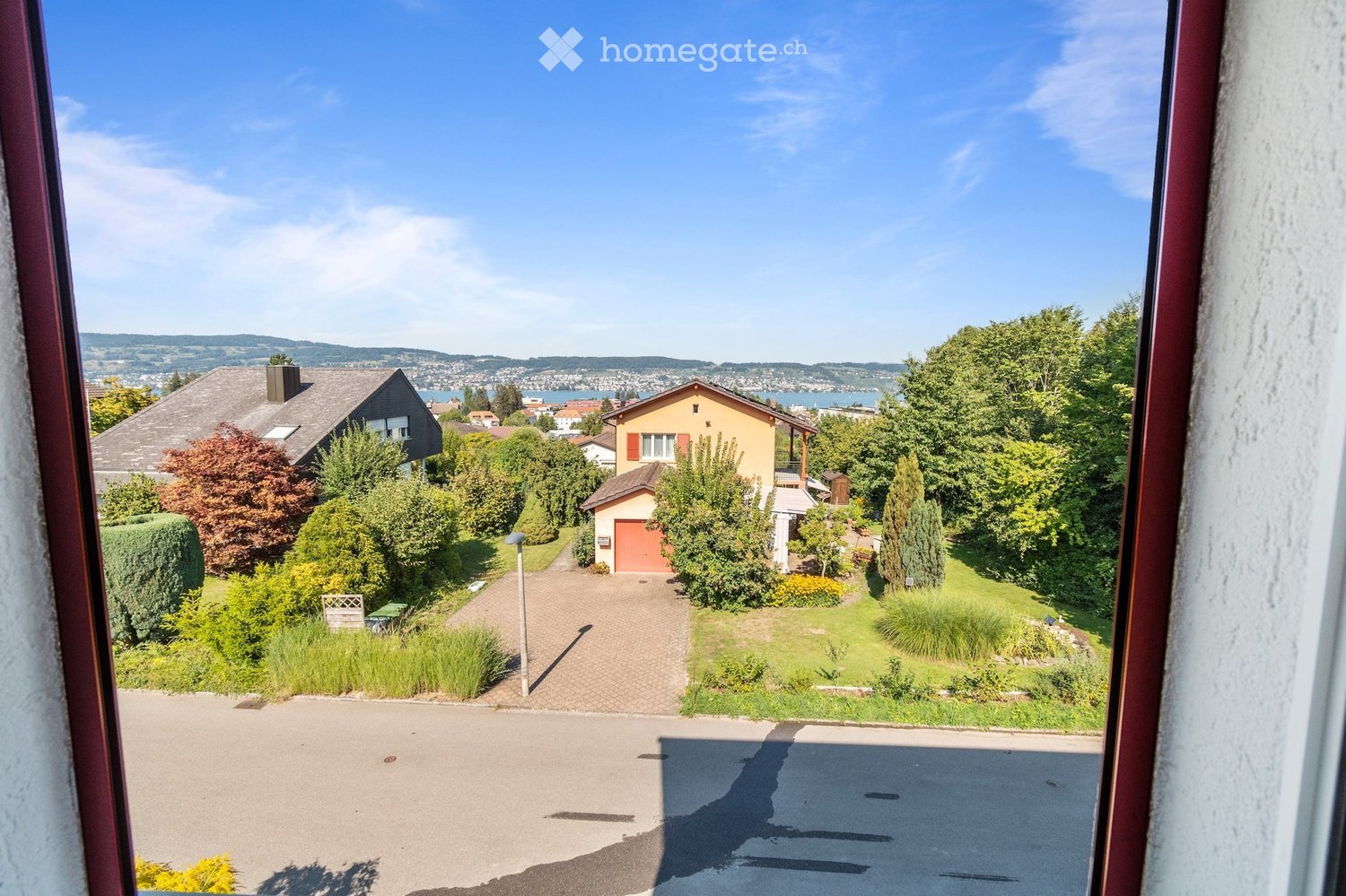 View from a window showing a suburban neighborhood with houses, gardens, and a scenic lake view in the background.