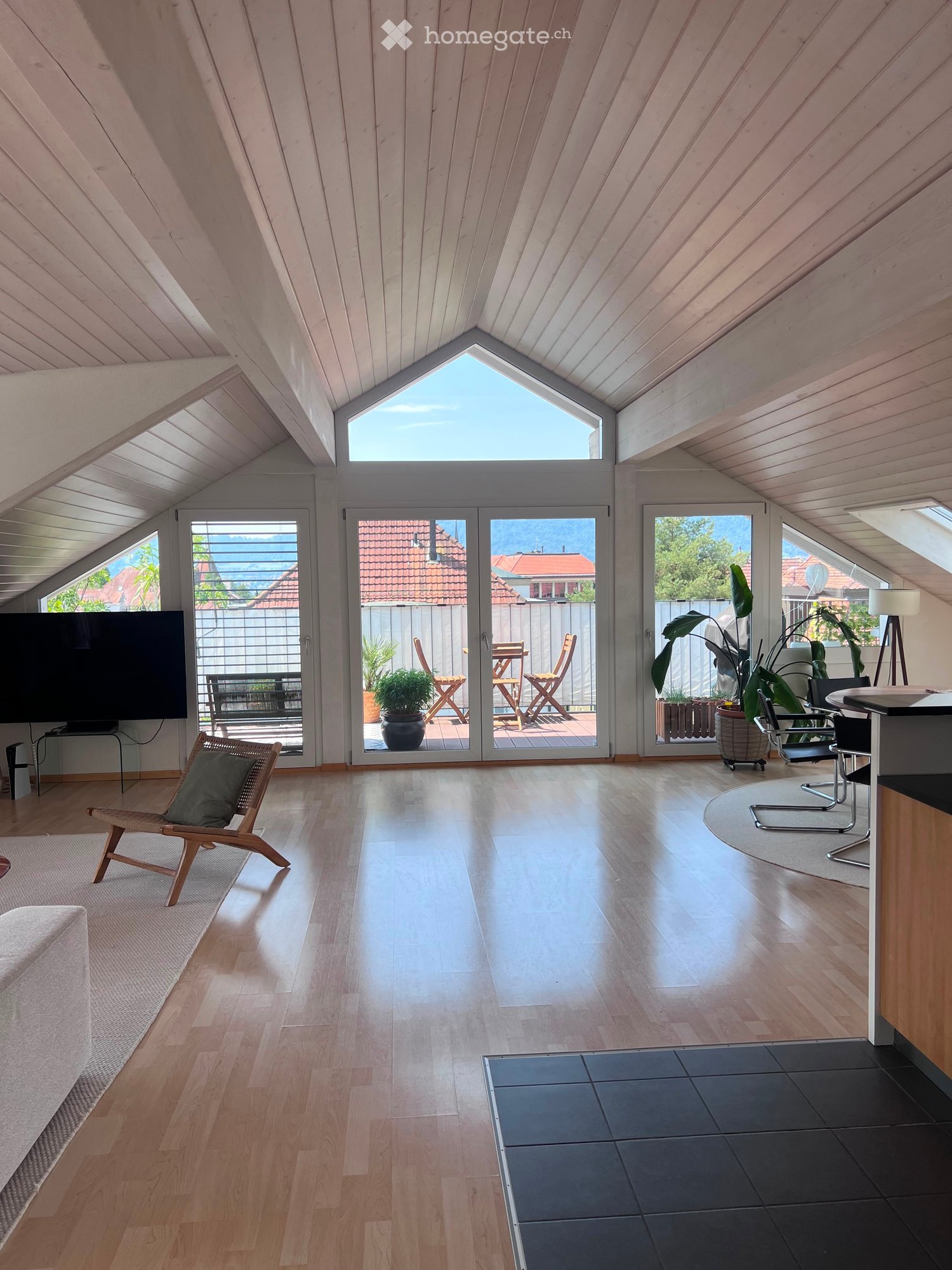 Bright, airy room with wooden floors, large windows, and a view of the outdoor area. Features a sofa, chair, table, television, and potted plants.