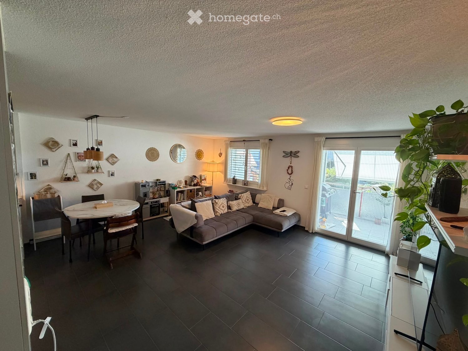 Modern living room with a grey sectional sofa, dining table with chairs, a lamp, and decorative mirrors on the wall. Large windows and a sliding glass door lead to a balcony.