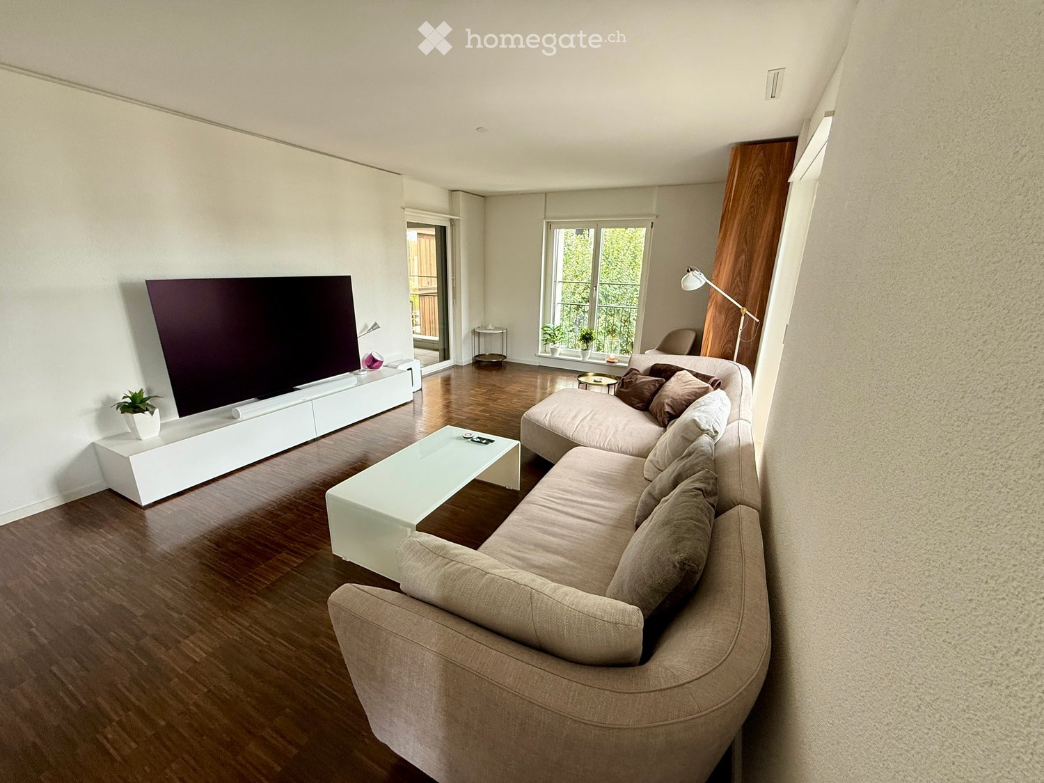 Modern living room with a large sectional sofa, coffee table, flat-screen TV on a white console, and a floor lamp near a large window with a view of a balcony.