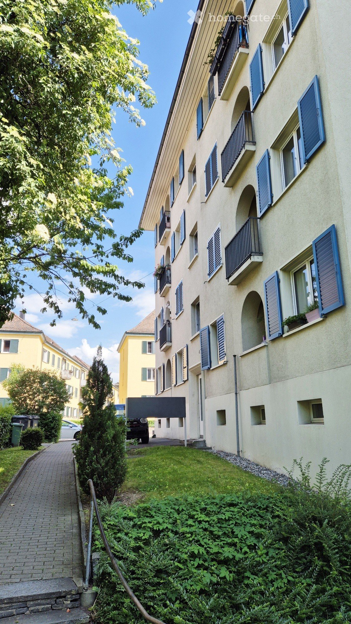 Multi-story residential building with light yellow facade, blue shutters, and multiple balconies. The building is surrounded by greenery and a paved pathway.