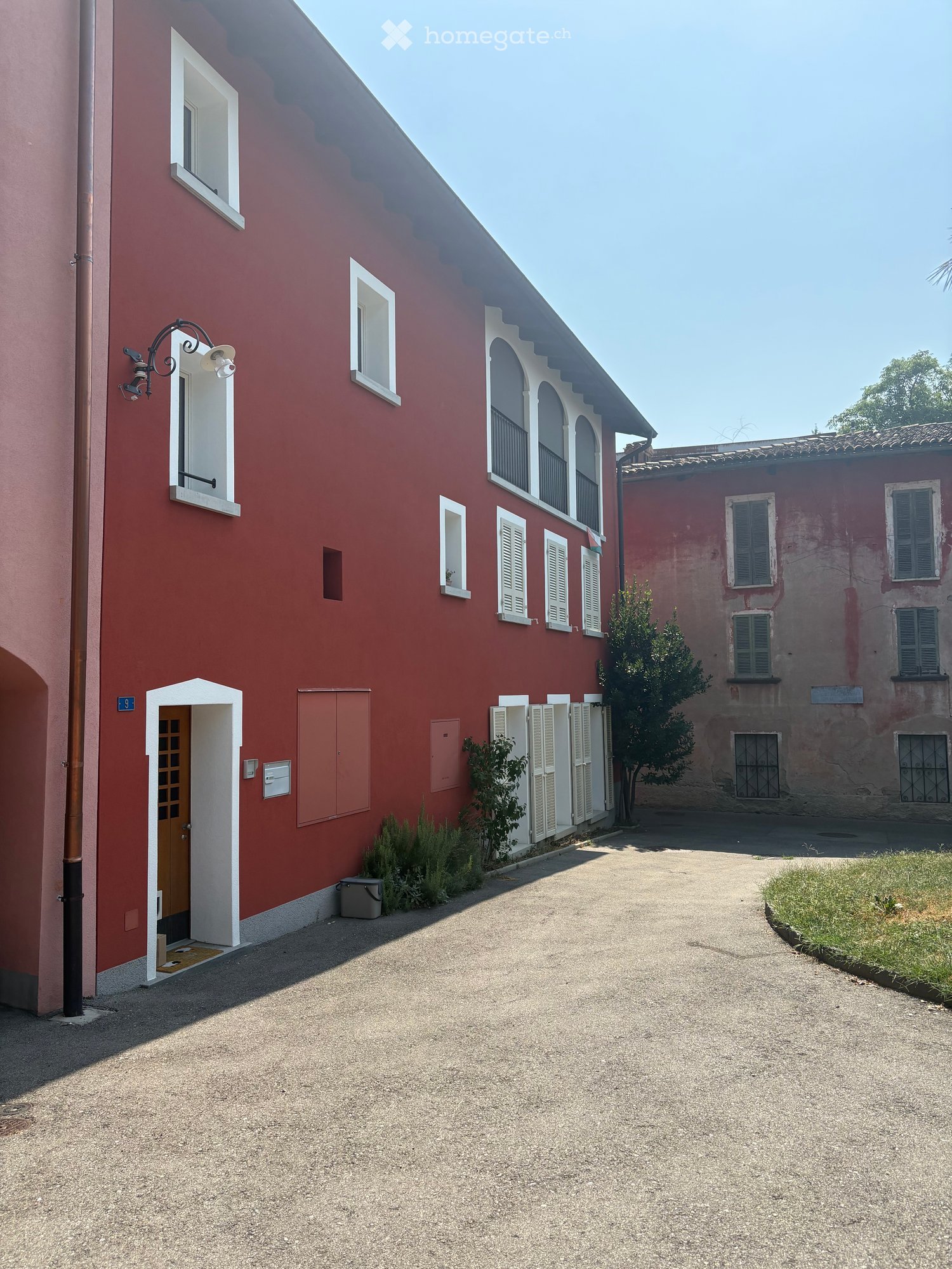 Red brick building with white-framed windows and shutters, a small tree, and a gravel driveway.