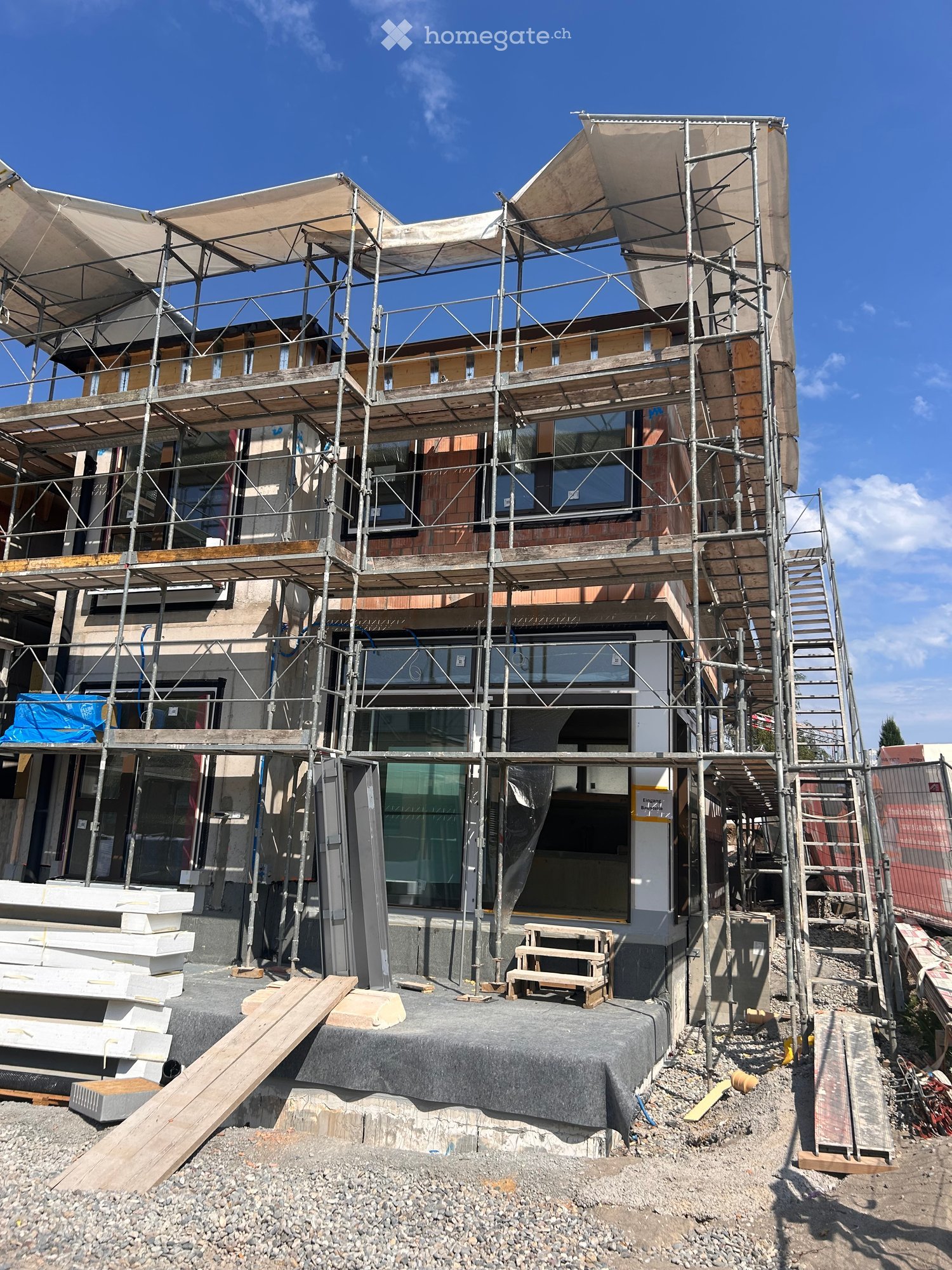 A modern house under construction with scaffolding, exposed wooden beams, and large windows. The structure features a raised ground floor and a contemporary design with visible construction materials and tools.