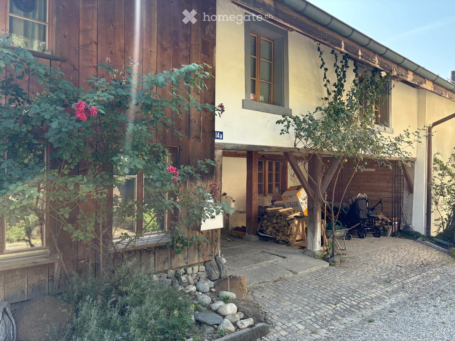 Two-story wooden house with a stone foundation, wooden shutters, and a small garden area with a stroller, firewood, and plants.