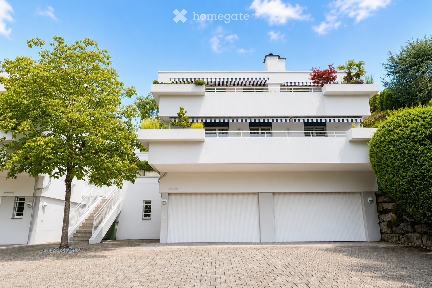Modern two-story white house with large garage, raised ground floor, and balcony terrace with greenery.