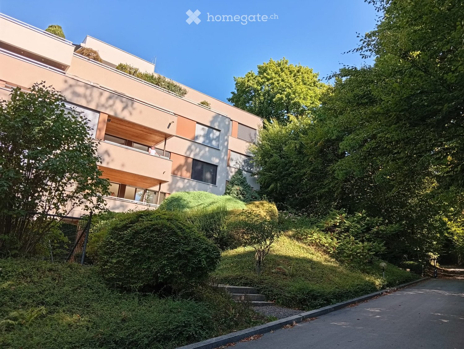 Modern multi-story building with balconies and large windows, surrounded by lush greenery and trees.