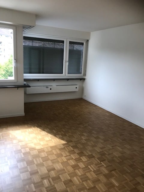 Empty room with wooden parquet flooring, large windows with blinds, and white walls.