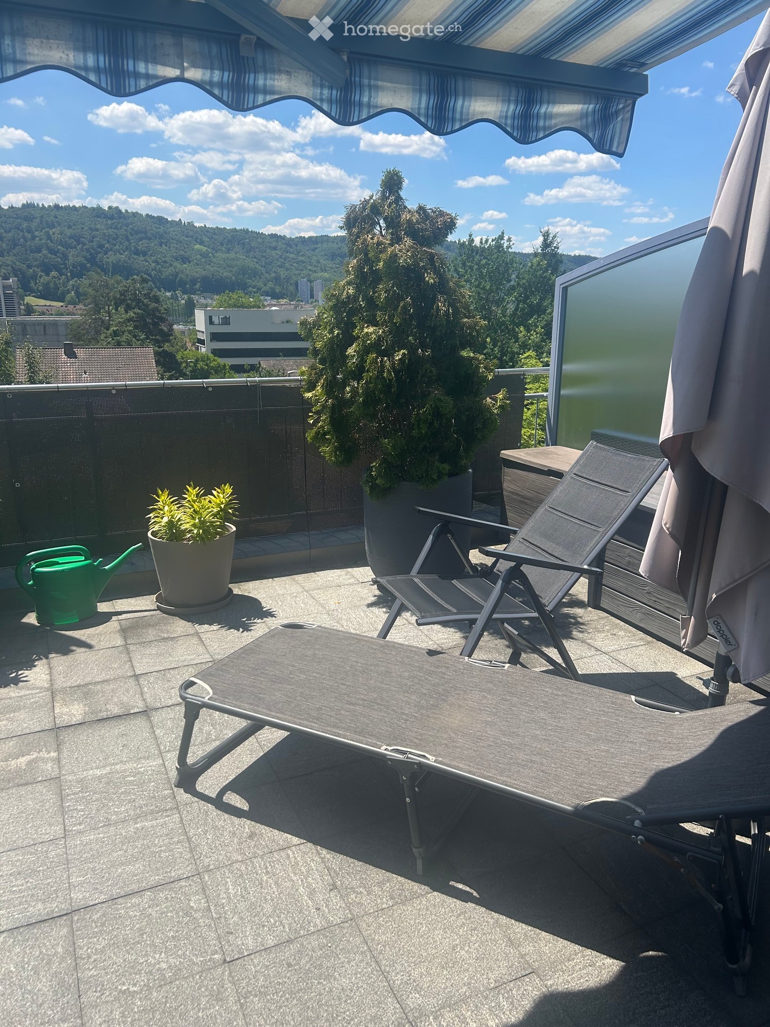 Balcony with lounge chairs, small table, umbrella, potted plants, and a scenic view of the city and hills.