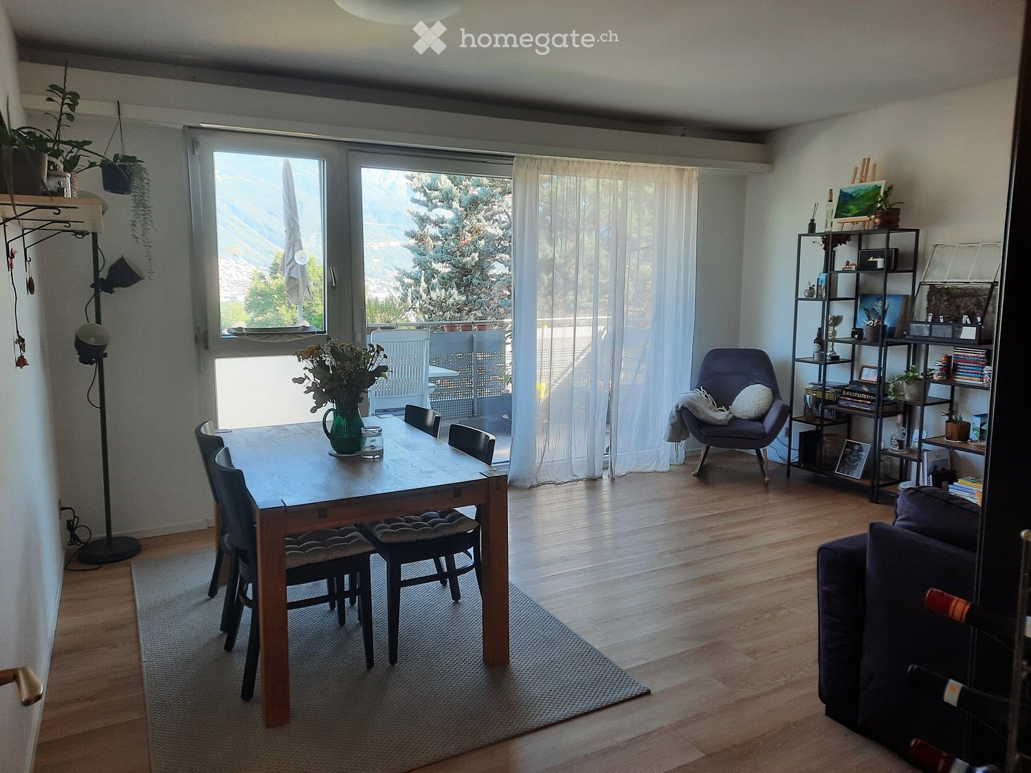 Bright dining area with wooden table, chairs, sofa, shelf, and view of greenery through large windows.