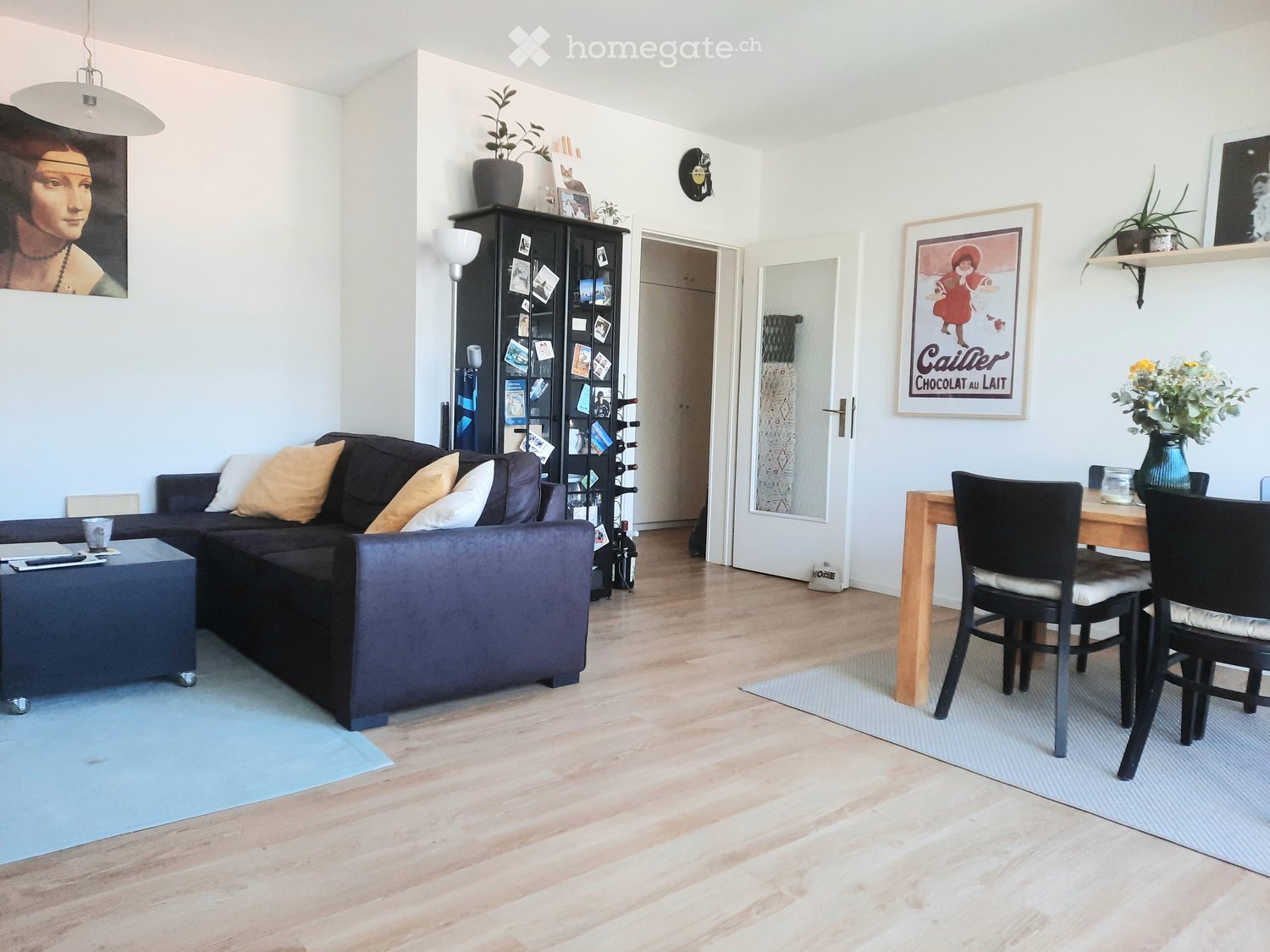 Modern living room with a dark grey sectional sofa, coffee table, dining table with chairs, a cabinet, and decorative items including artwork and plants.