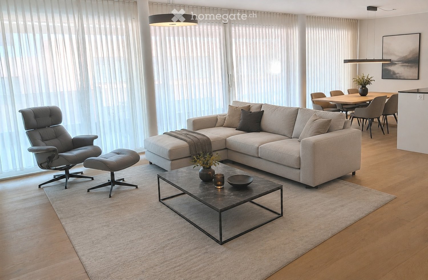 Modern living room with a grey sectional sofa, leather armchair, coffee table, dining area with chairs and table, pendant lights, and large windows with sheer curtains.