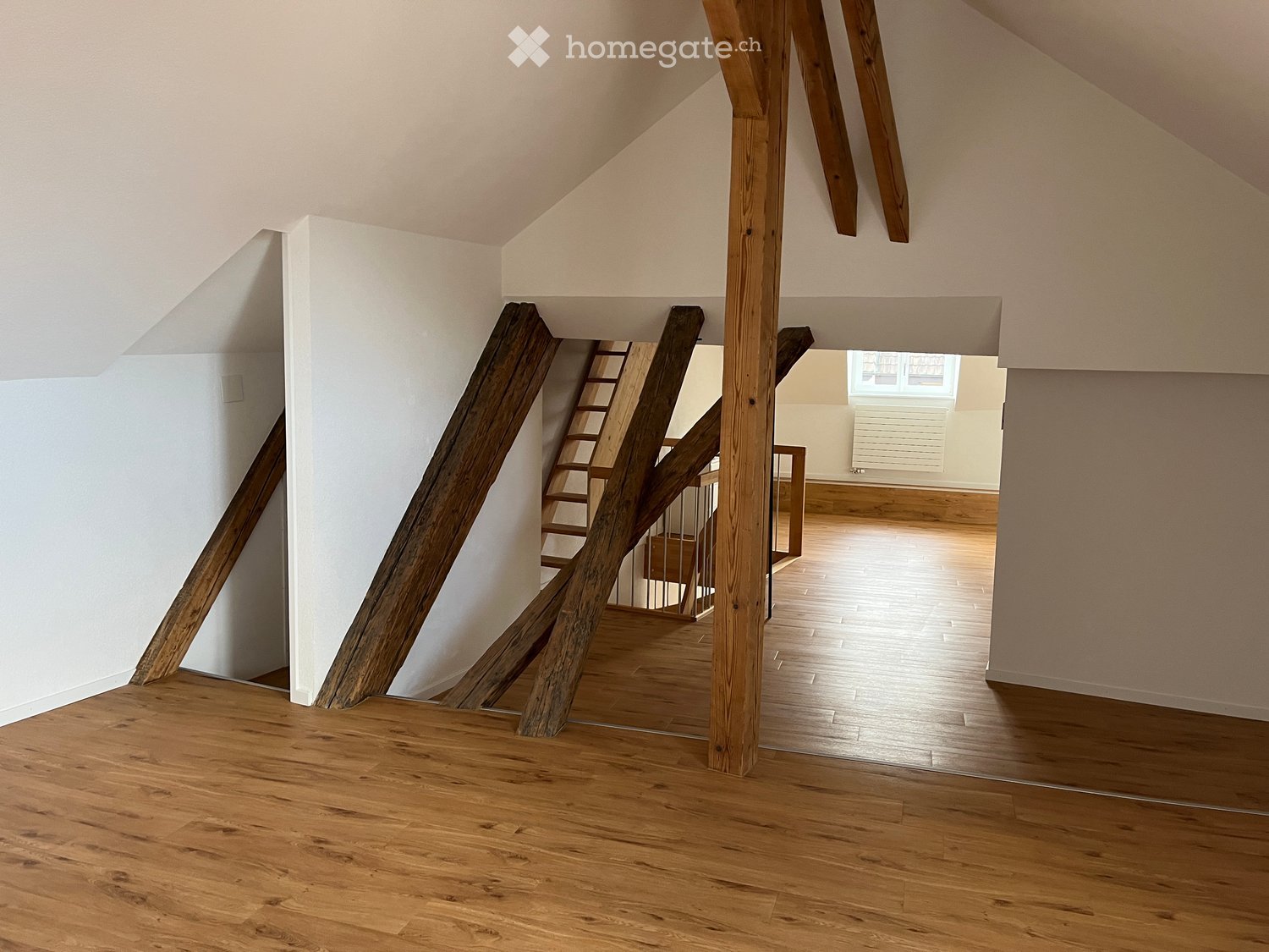 Empty hallway with wooden floors, white walls, and exposed wooden beams. A staircase leads to another room with wooden flooring and white walls.