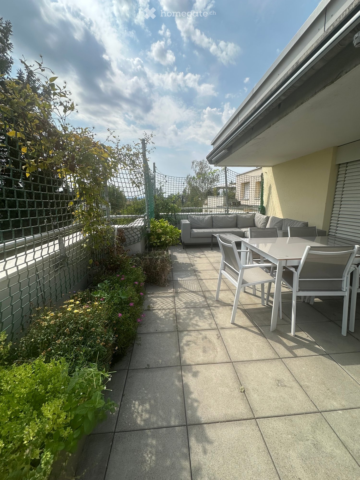 Balcony with tiled floor, sofa, table, chairs, and plants along the edge.