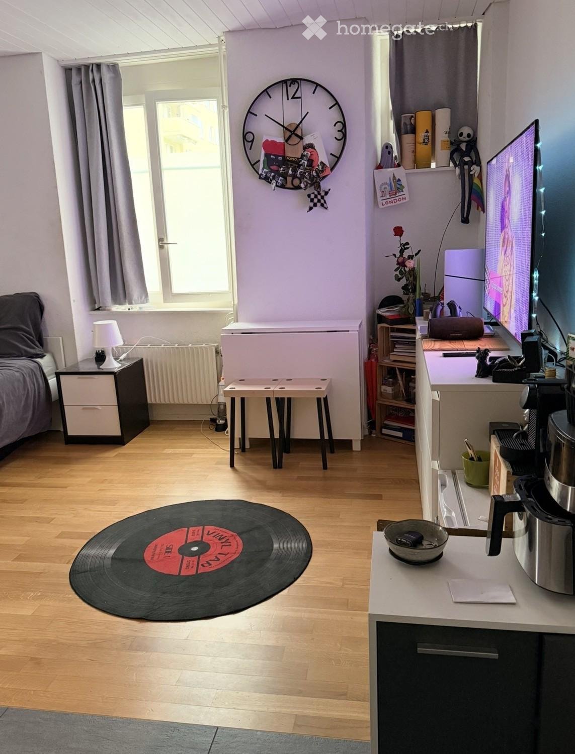 A modern, well-lit room with a bed, nightstand, lamp, two stools, a large vinyl record as a rug, a desk with a screen, and a small kitchen area with a fridge, pot, and bowl.