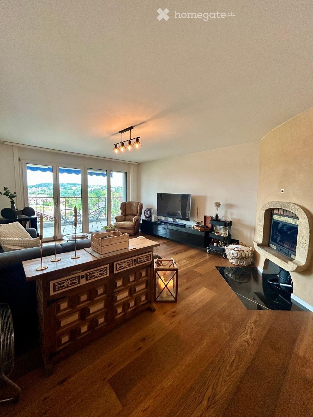Living room with hardwood floors, large windows, balcony access, fireplace, sofa, armchair, coffee table, TV, and decorative items.