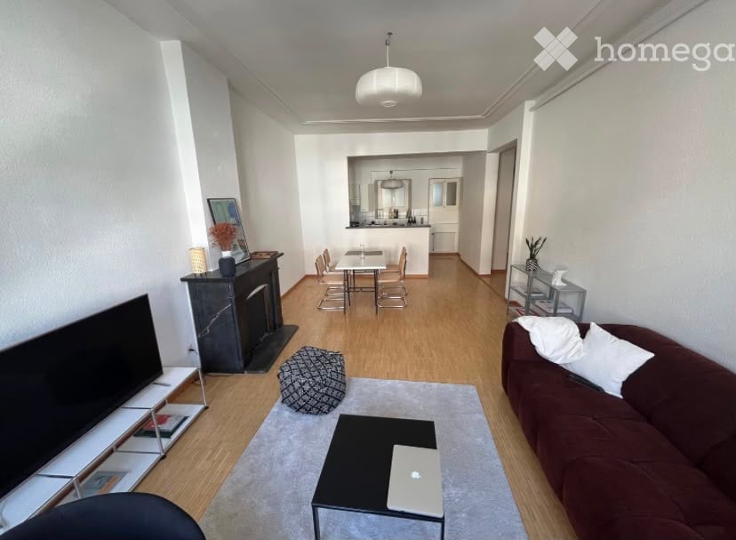 Modern living room with a brown sofa, coffee table, laptop, ottoman, fireplace, and dining area with chairs and a table. The room features light wooden flooring and white walls.
