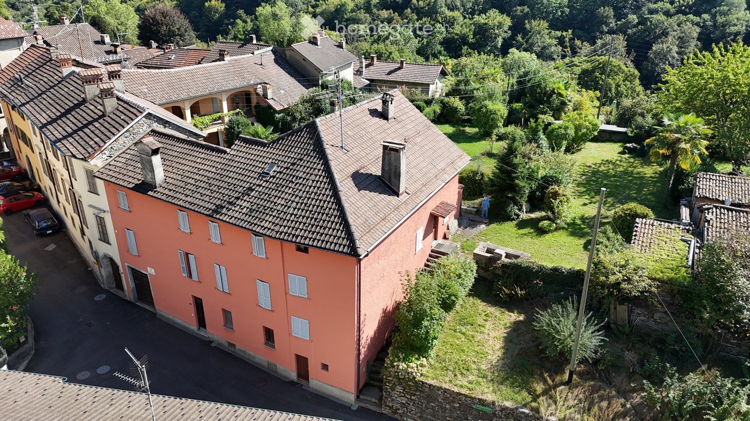 Aerial view of a multi-story residential building with a terracotta roof, surrounded by greenery and neighboring houses.