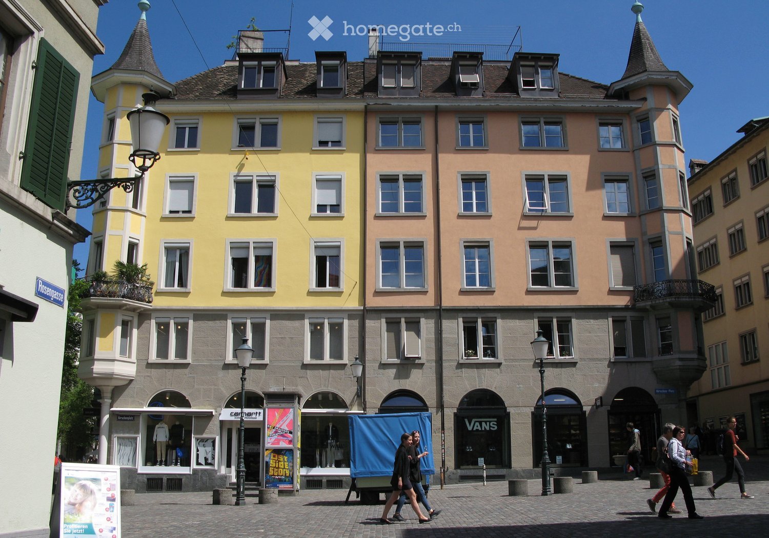 A multi-story building with a yellow and pink facade, featuring multiple windows, balconies, and shops at the ground level. The building has ornate architectural details, including spires and decorative elements.