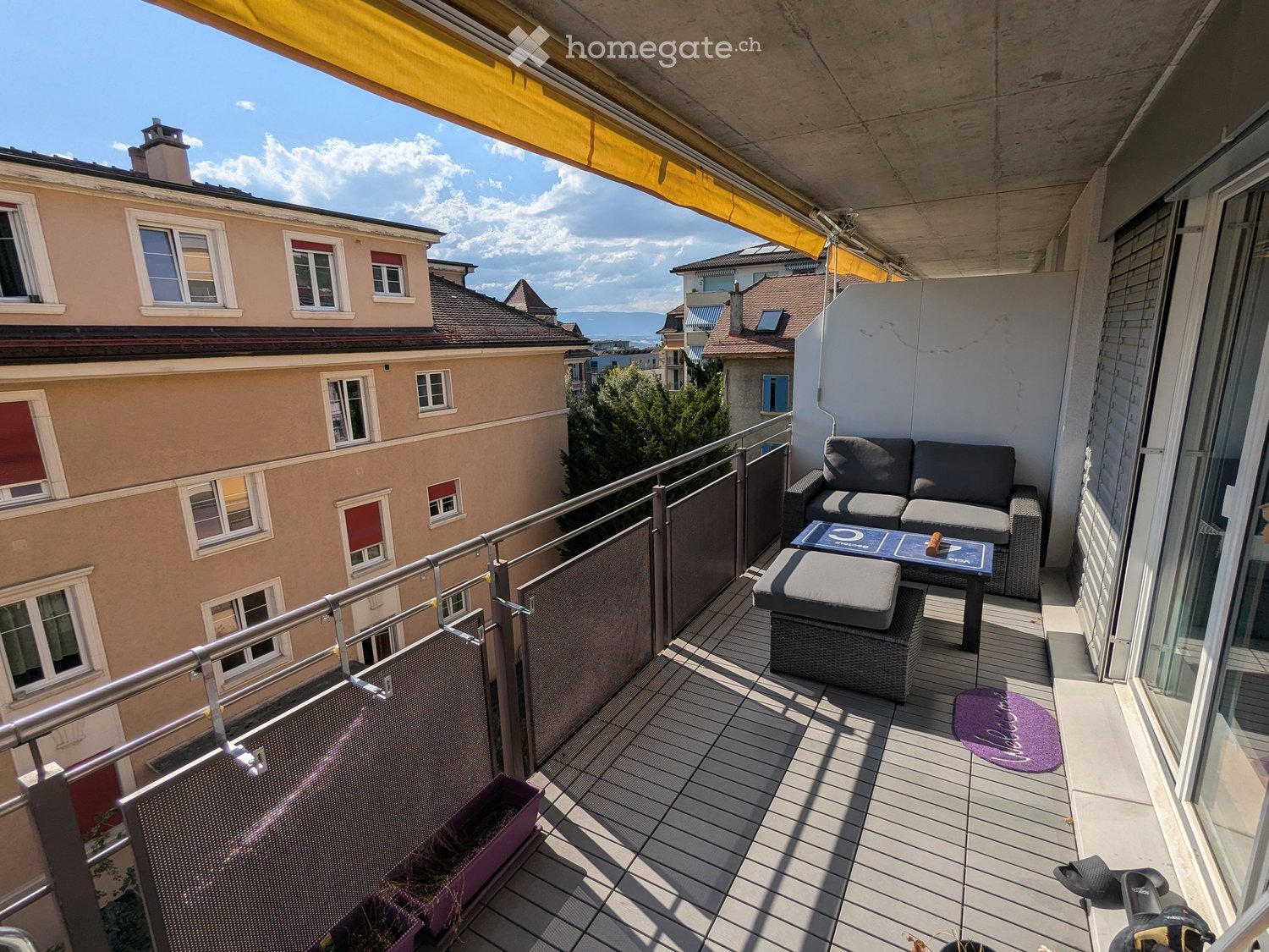 Balcony with wooden decking, metal railing, sofa, table, stool, rug, and bicycle.