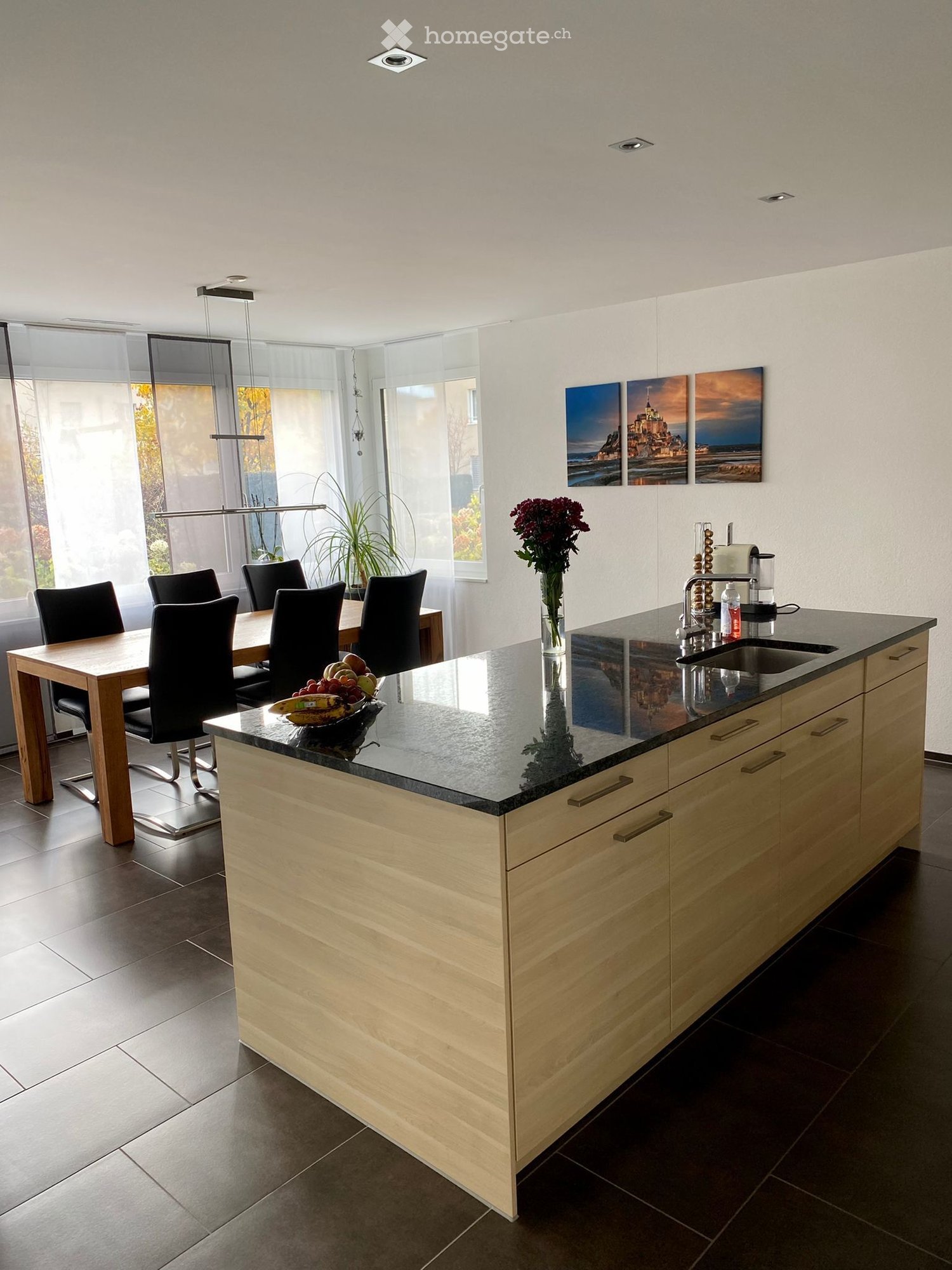 Modern kitchen with a light wood island, black granite countertop, sink, and a dining area with a wooden table and black chairs. There is a vase with flowers on the island and a bowl of fruit on the table.