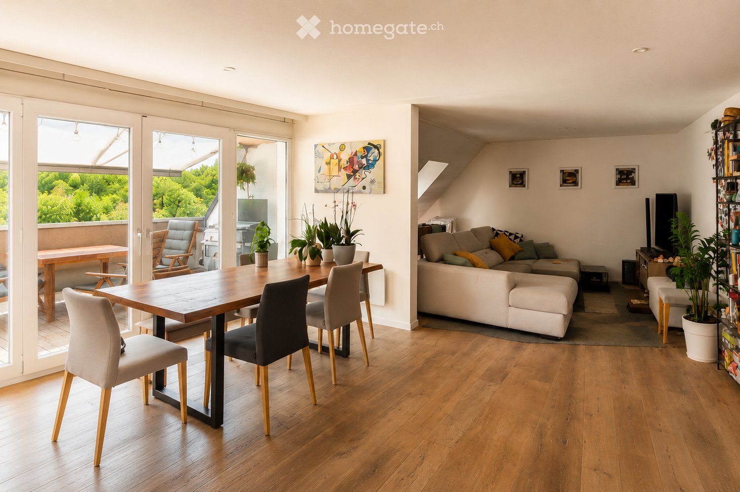 Bright, open-plan living and dining area with wooden flooring, large windows, and a balcony. Features a white sectional sofa with cushions, a coffee table, dining table with chairs, potted plants, and a TV on a stand.