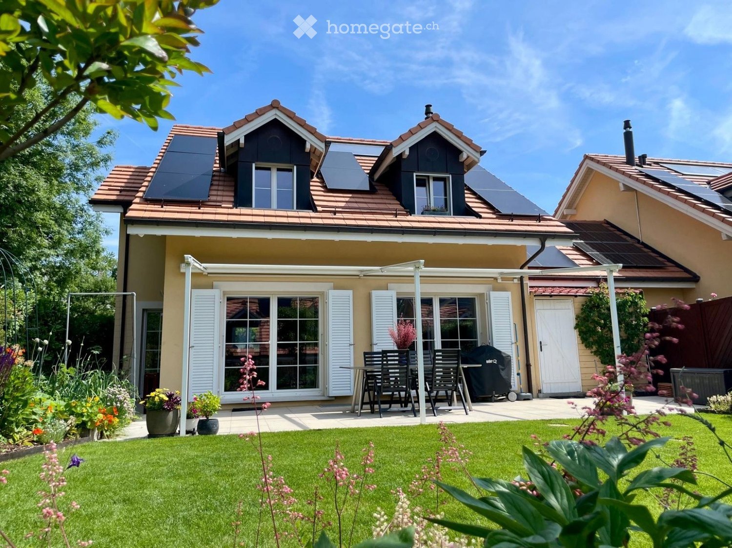 A well-maintained garden with a lawn, flower beds, and a patio area with a table, chairs, and a grill. The house features solar panels on the roof and large windows with shutters.