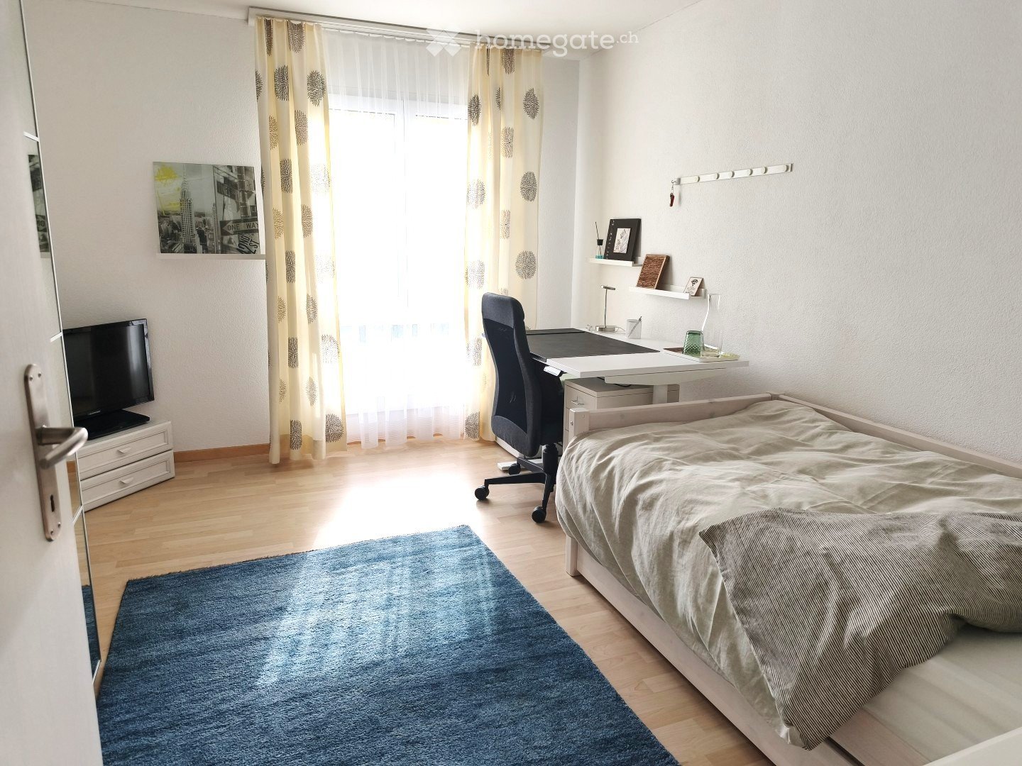 A well-lit bedroom with a bed, desk, chair, and a TV. The room features a large window with curtains, a blue rug, and a shelf with decorative items.