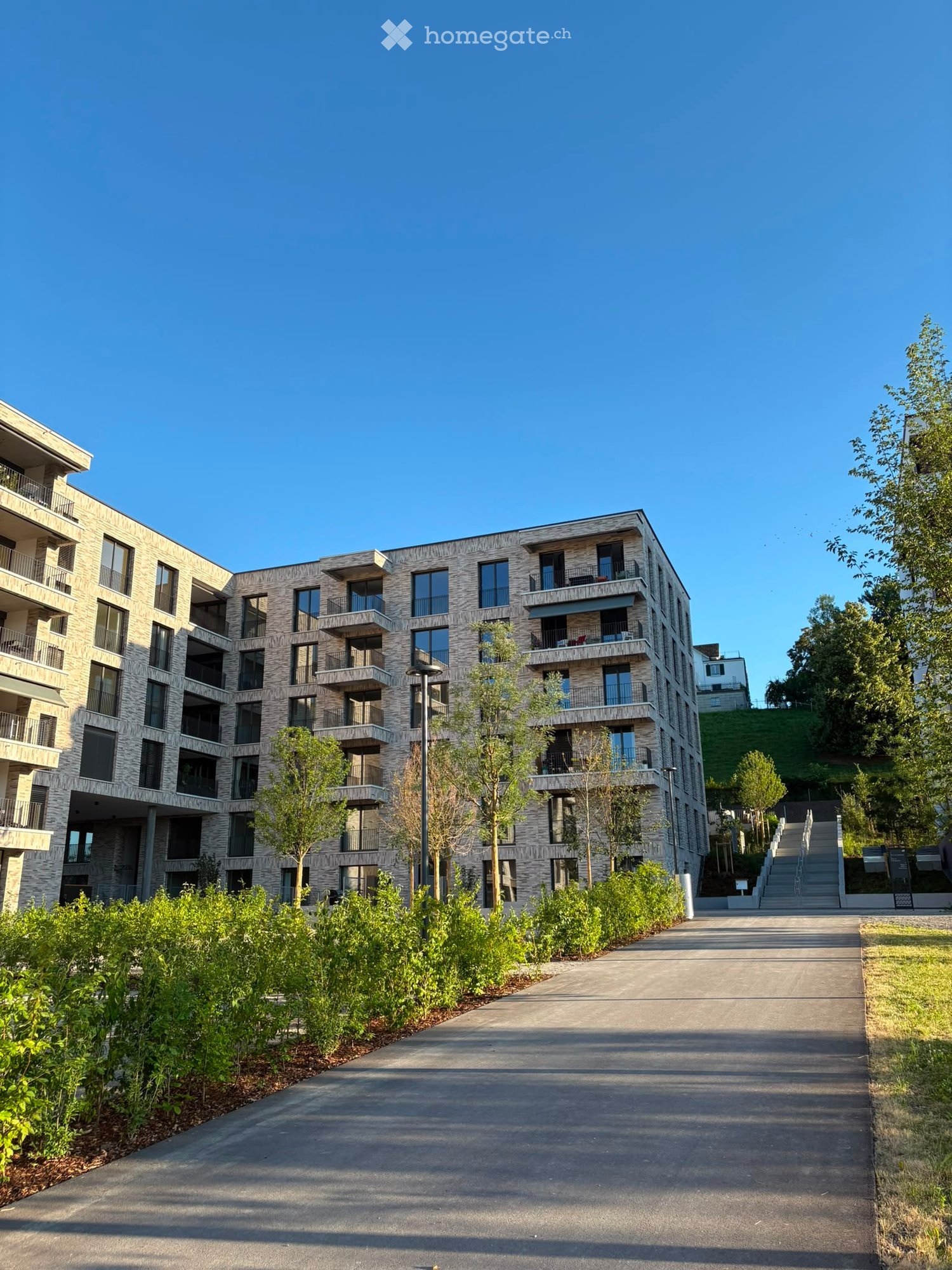 Modern multi-story residential building with balconies and large windows, surrounded by greenery and a paved walkway.