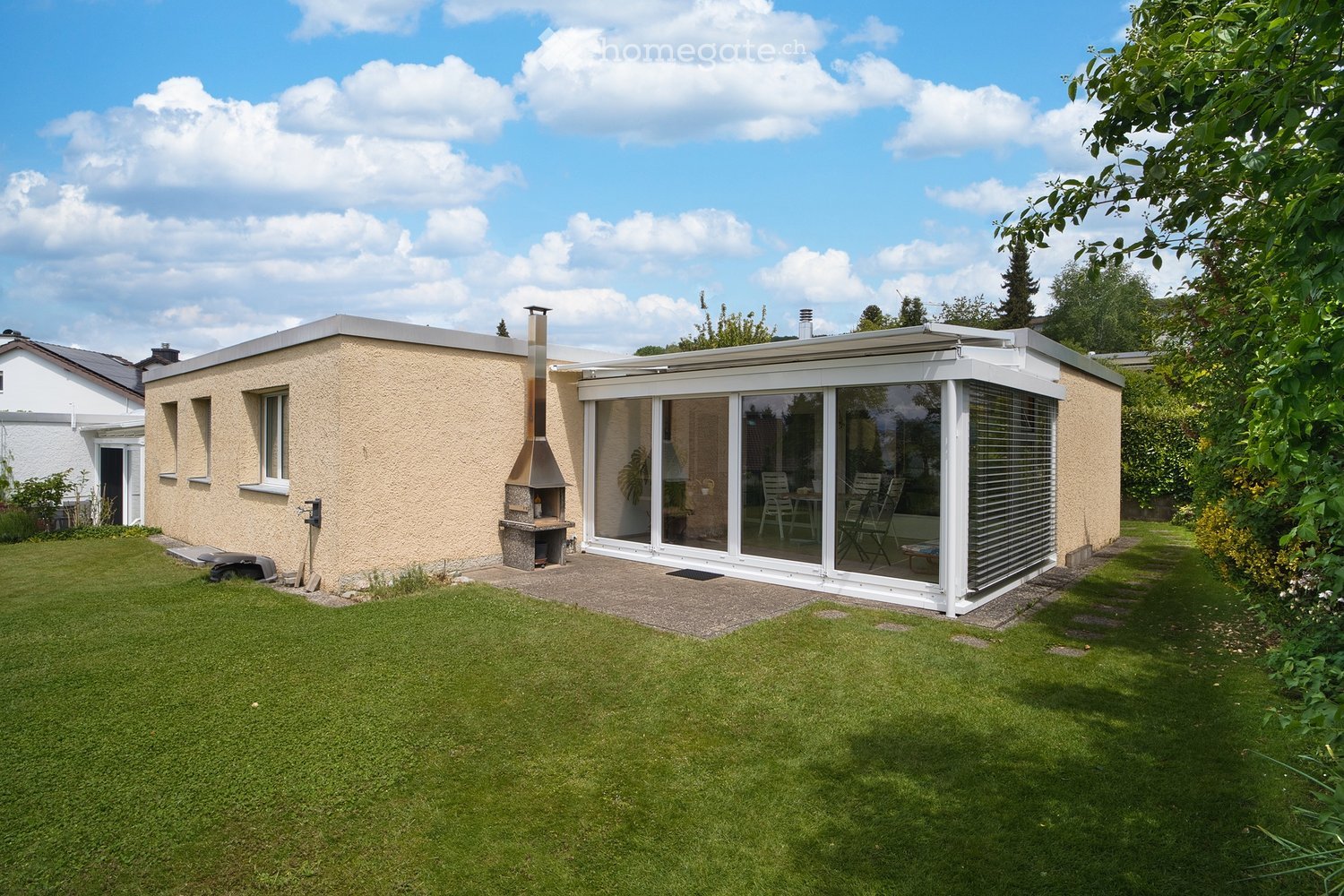 A modern single-story house with a flat roof, beige exterior, and a large glass-enclosed patio area. The patio contains outdoor furniture, including chairs and a table. The surrounding area features a well-maintained lawn and trees.