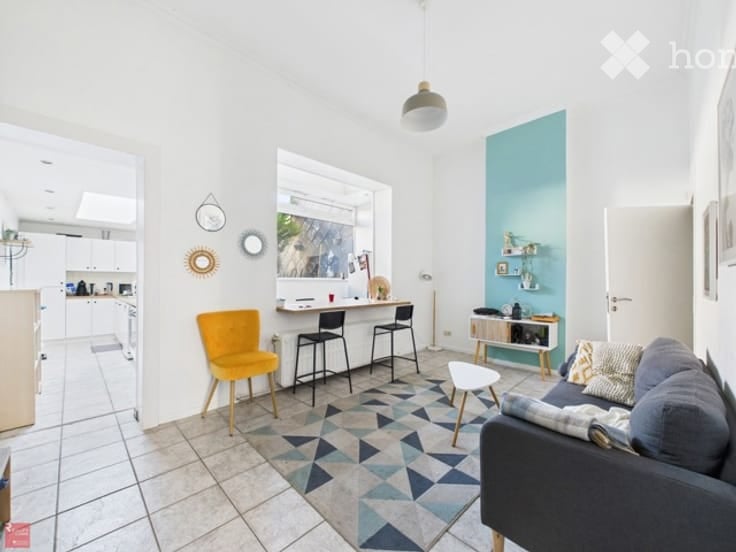 Bright, modern living space with a grey sofa, coffee table, yellow accent chair, and small kitchen area with bar stools. Features a geometric rug, white walls, and a blue accent wall.