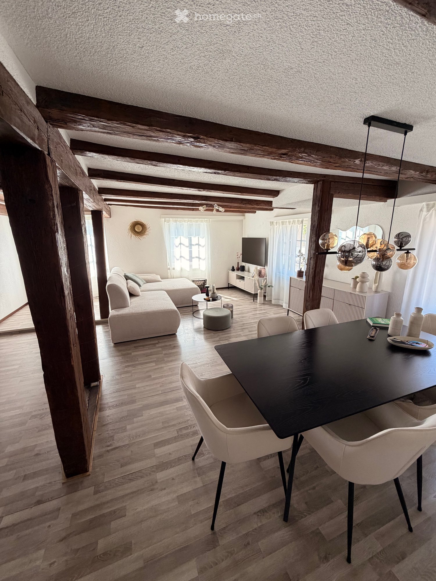 Open-concept living and dining area with exposed wooden beams, hardwood flooring, a sectional sofa, a coffee table, an ottoman, a dining table with chairs, a television, and decorative vases.