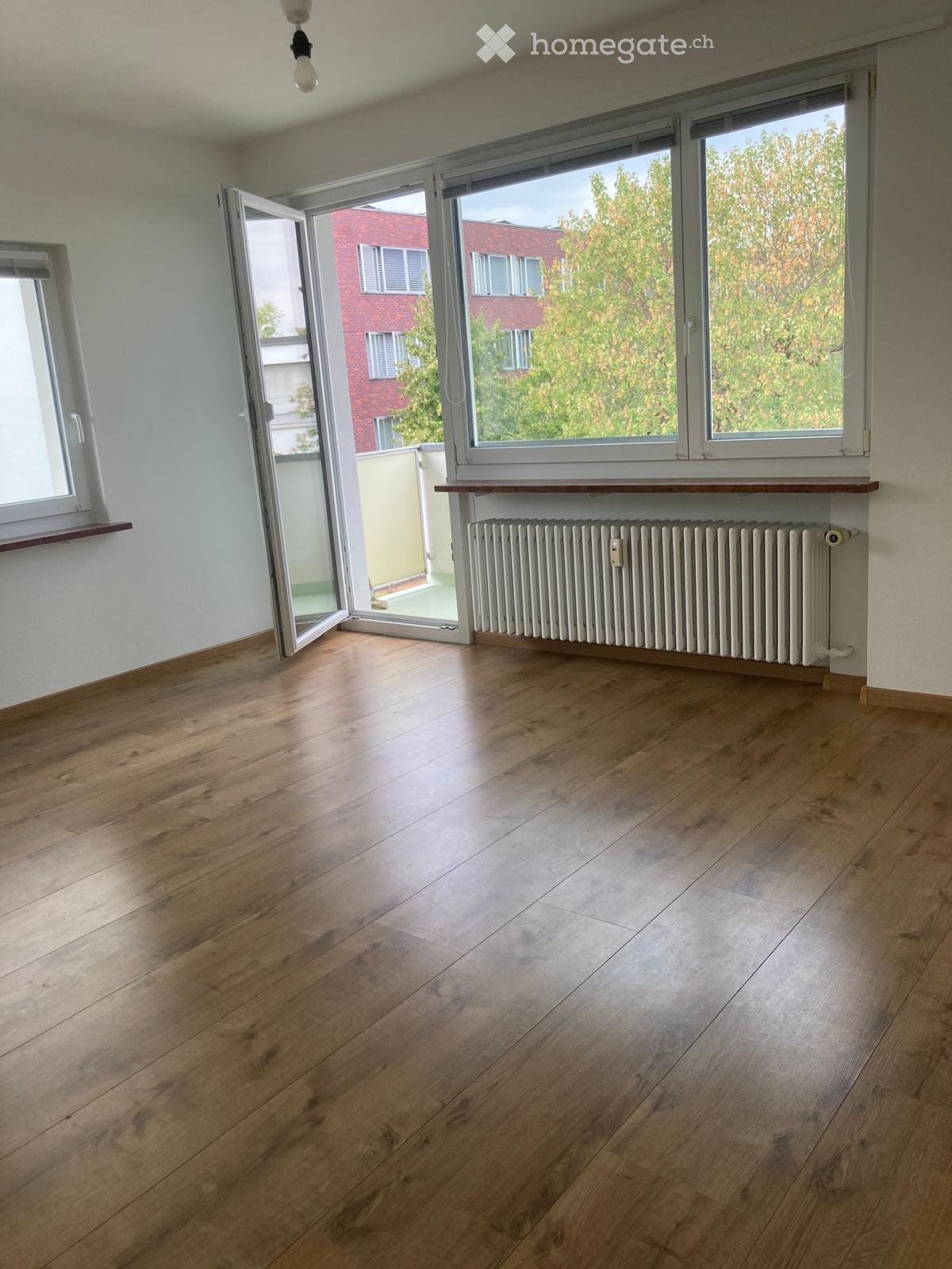 Empty room with wooden flooring, large windows, radiator, and open sliding doors leading to a balcony.