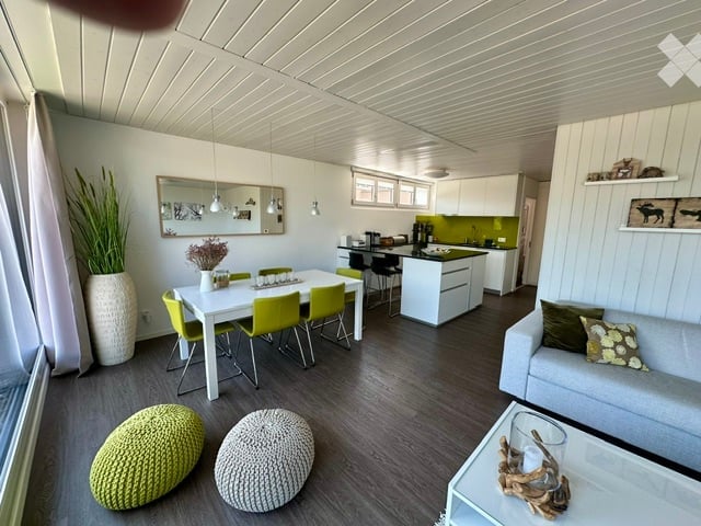 Open-plan living and dining area with modern furniture, including a grey sofa, coffee table, dining table with yellow chairs, and kitchen cabinets. Decorative elements include a large mirror, vase with plants, and pendant lights.