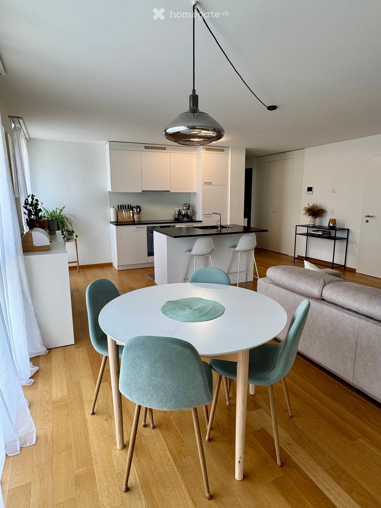 Modern open-plan living and dining area with a round white dining table, four light blue chairs, a grey sofa, kitchen island with white cabinets, and pendant lighting.